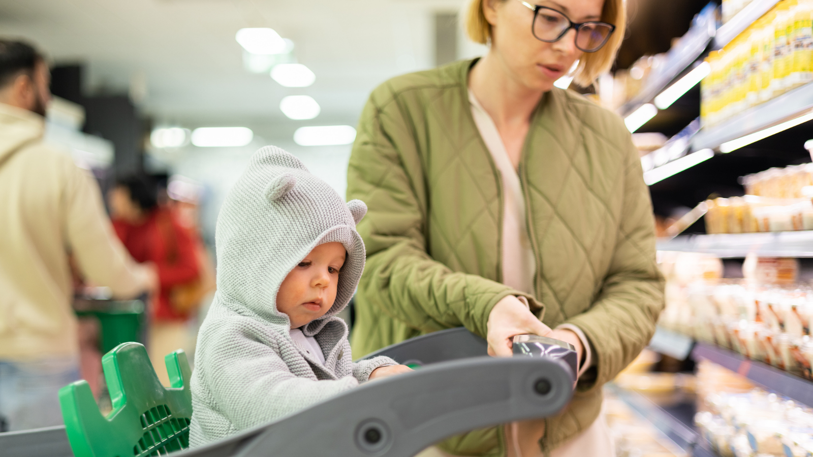 Bebê sentado no carrinho de compras de um supermercado ao lado da mãe