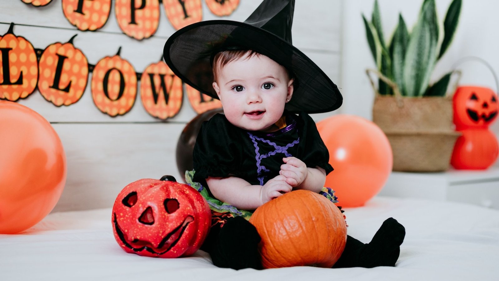 Bebê fantasiada de bruxinha, cercada de abóboras decorativas de Halloween