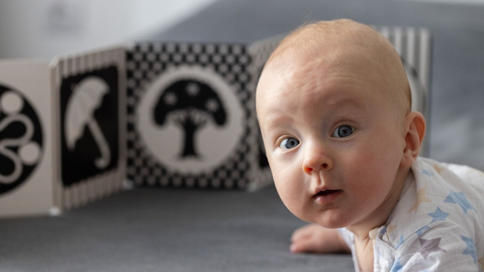 Bebê de bruços olhando para a frente, com um painel de imagens em preto e branco desfocado ao fundo
