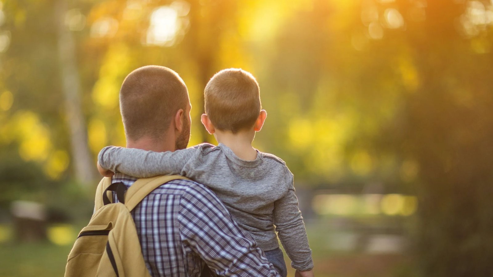 Pai de costas carregando o filho pequeno no colo, com uma mochila nas costas, em um parque ao entardecer