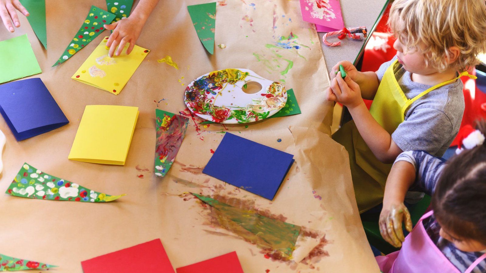 Crianças pintando árvores de Natal de papel e cartões coloridos com tinta, em uma mesa forrada de papel kraft na sala de aula
