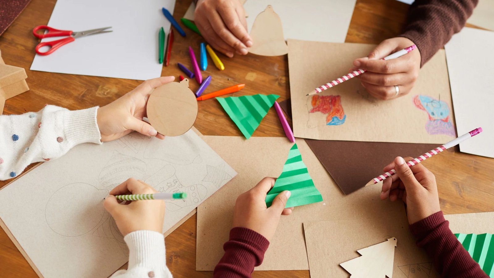Mãos de crianças e adultos fazendo artes de Natal com papel dobrado, enfeites de madeira e lápis de cor
