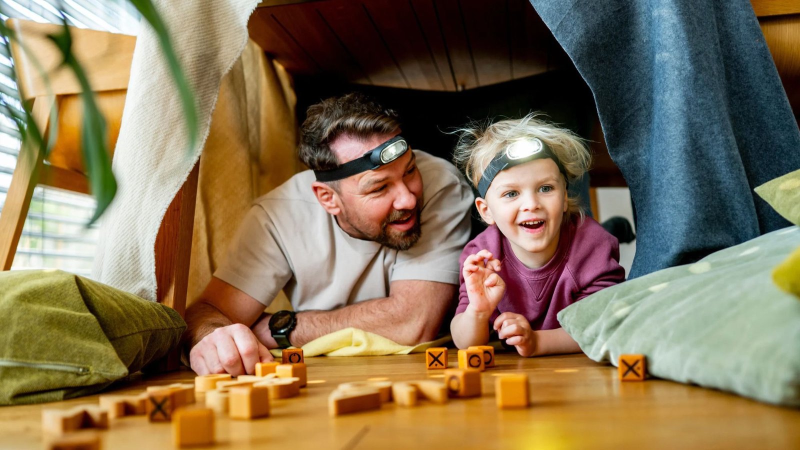 Pai e filho brincando dentro de uma cabana de lençol, com lanternas de cabeça e blocos de madeira, durante as férias